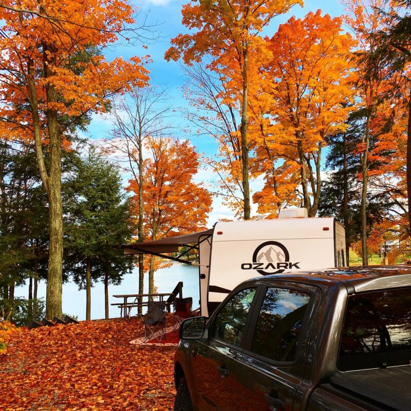 an rv and a black truck parked at a campground by the water surrounded by orange fall foliage