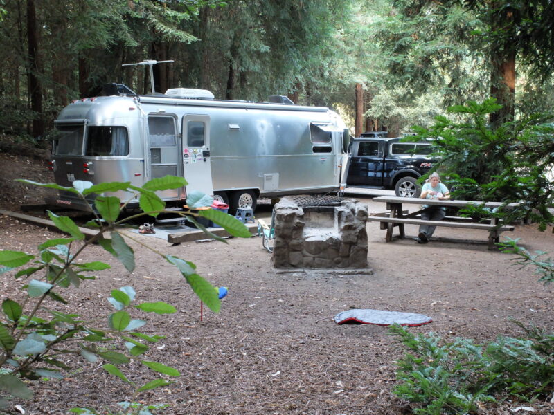 a person sits at a picnic table next to a silver airstream parked at a campsite