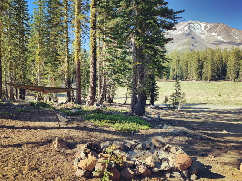 a campsite in the woods with a hammock, rock circled fire pit and snowy mountains in the background
