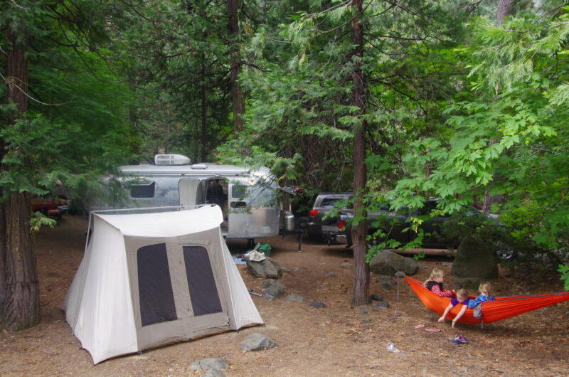 children sit in an orange hammock hung at a campground next to a pop up tent and airstream trailer