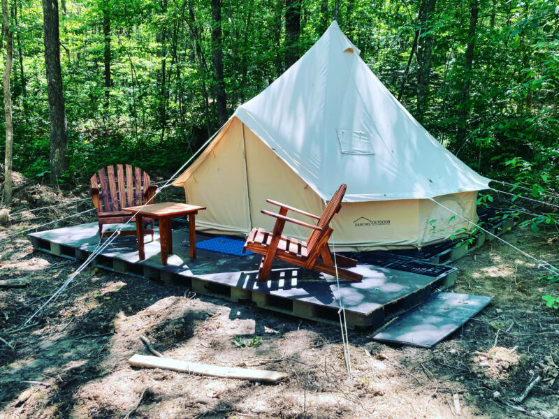 a campsite in the woods with a white glamping tent with a small patio and two adirondack chairs
