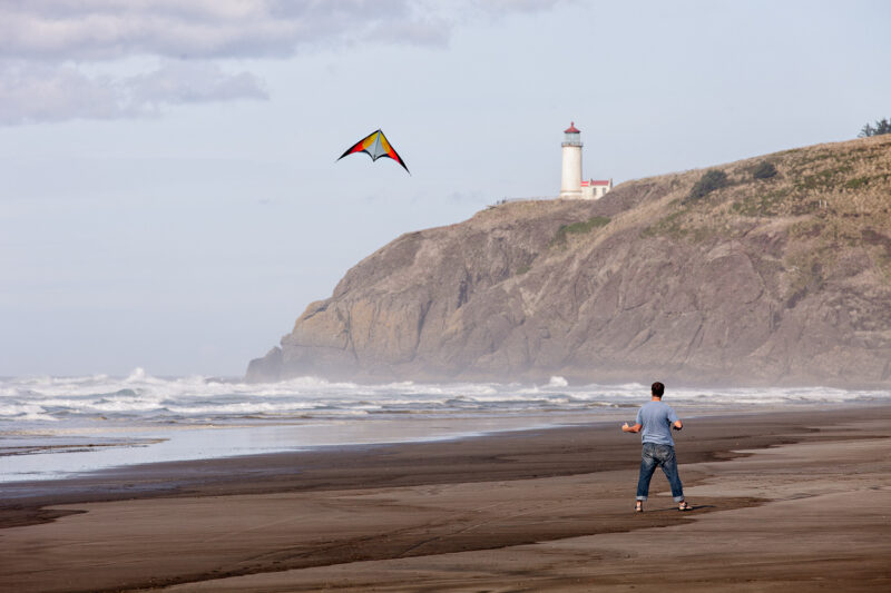 A person flies a kite on a misty beach while a lighthouse stands in the background.