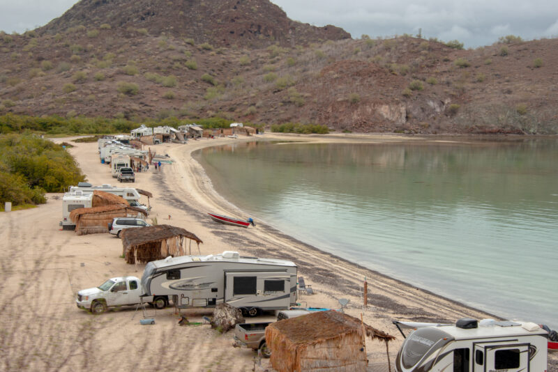 A variety of RVs sit on a sandy beach lining the shores of a large body of water.