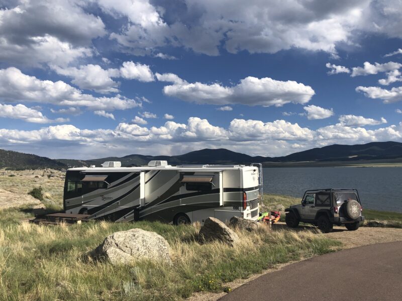 A motorhome sits in a scenic RV campground surrounded by water, brush, and hills.