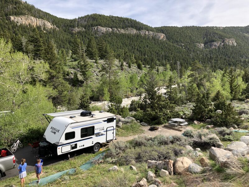 A travel trailer RV sits among a tree-covered landscape with mountains in the distance.