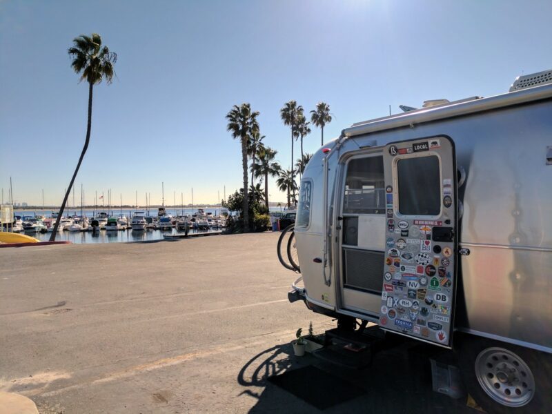 The door of an Airstream trailer is flung open, displaying a variety of travel-related stickers, as the camper looks out at an ocean setting filled with palm trees.