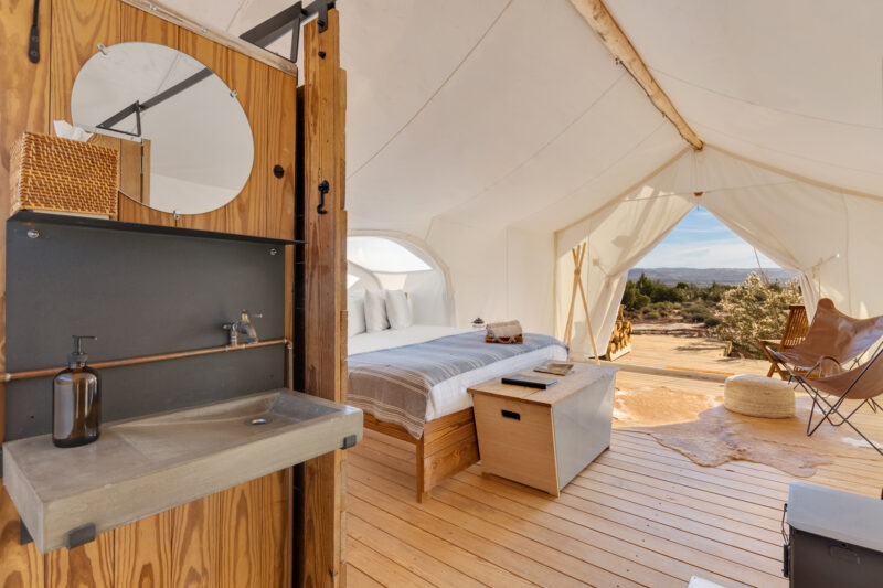 The inside of a glamping tent reveals a bed sporting fashionable, crisp linens and a sink.