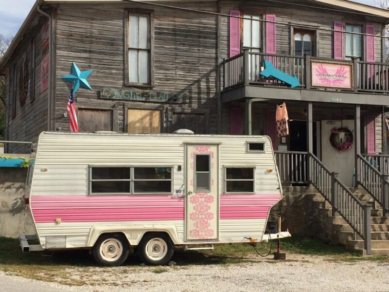 A retro pink and white travel trailer sits parked in front of a wood building