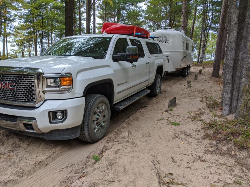 Truck towing a travel trailer parked at a campsite in a shaded wooded area with a sandy ground