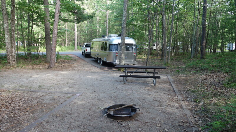 Silver Airstream trailer parked at a campsite in a heavily wooded area