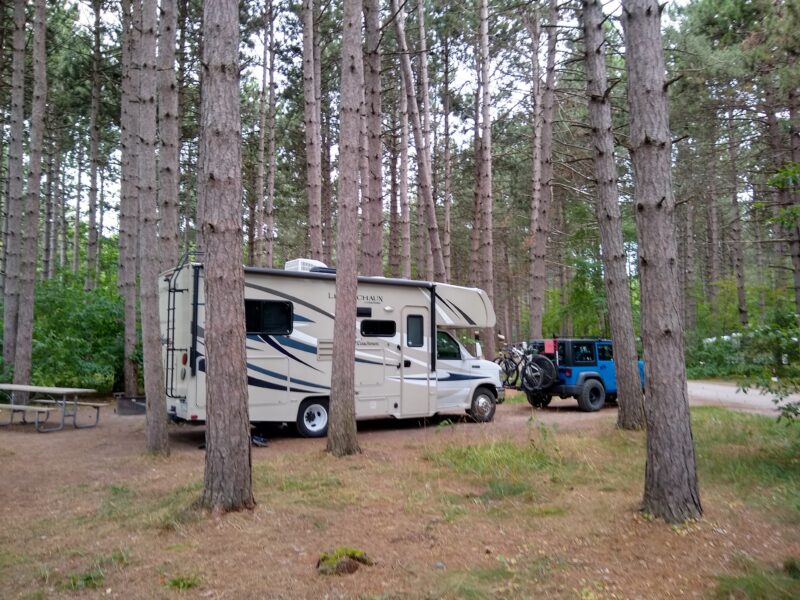 Class B motorhome parked in a wooded area with a blue Jeep parked in front of it