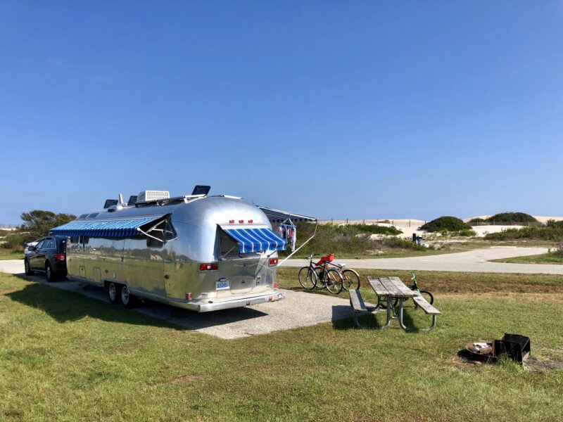 Silver RV parked at a beach campsite with rolling dunes in the background
