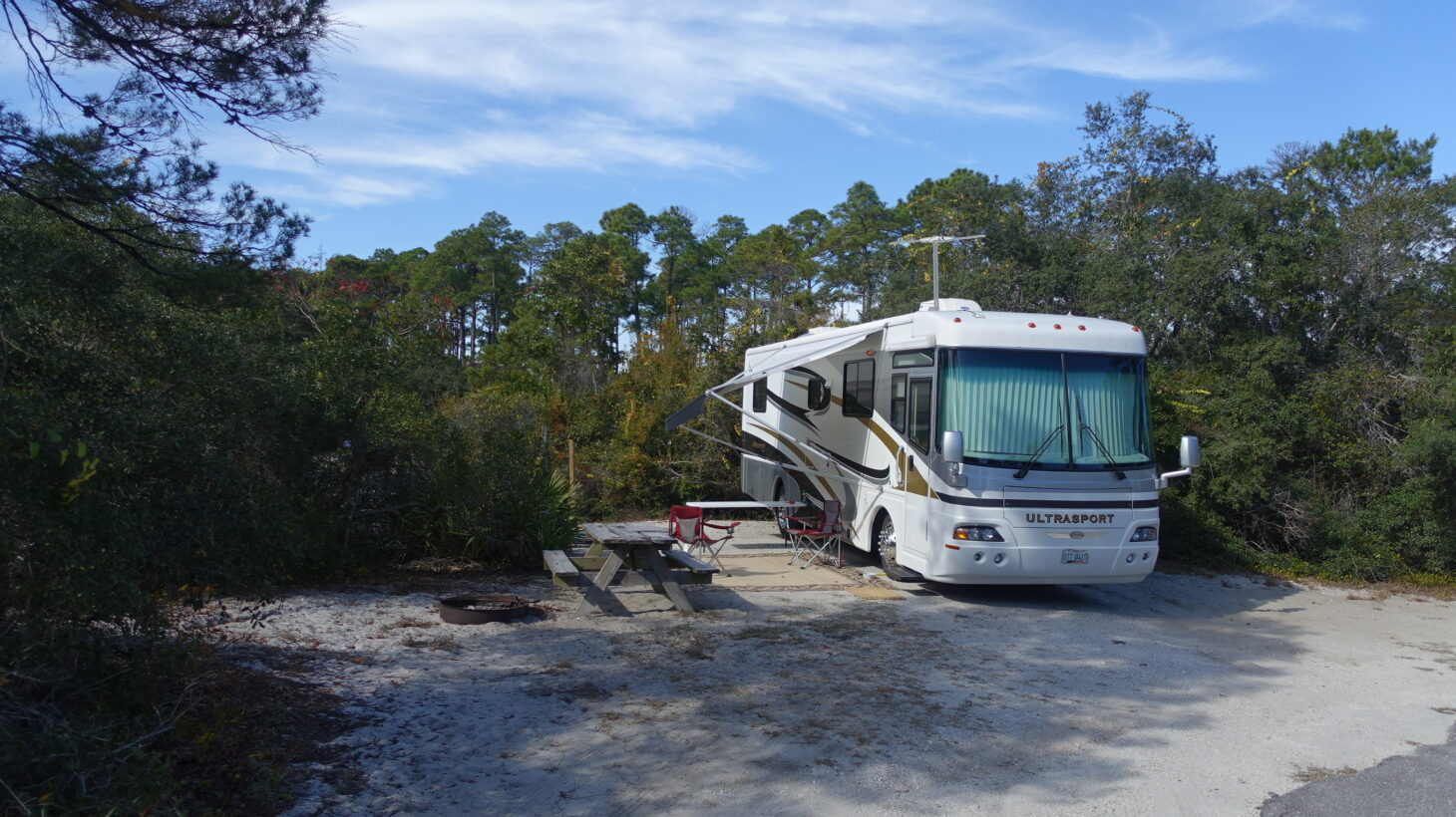 Large motorhome parked at a sandy campsite with a wooded area behind it