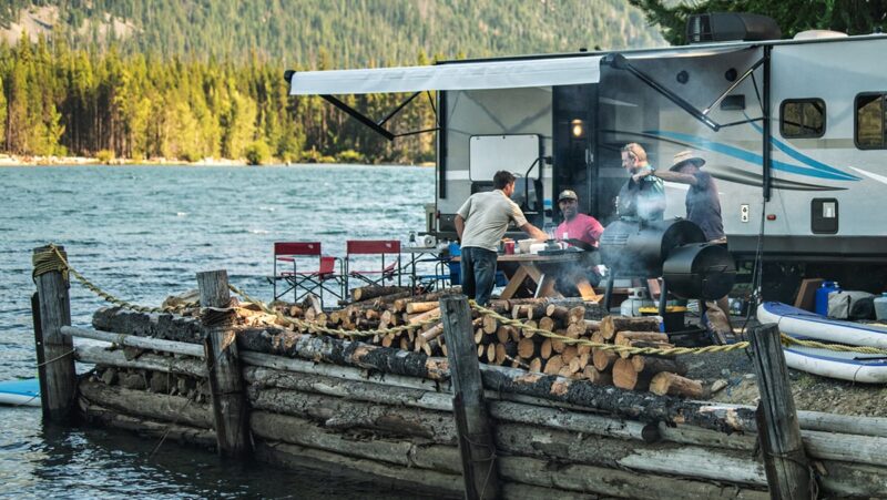 Group of people cooking and eating outside of an RV next to a lake