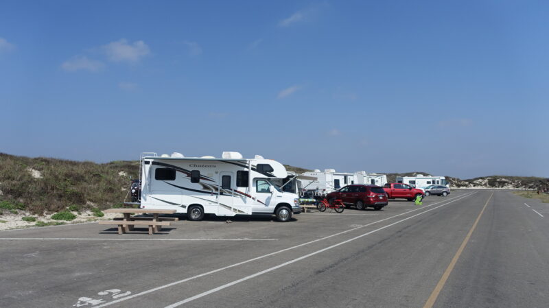 Multiple RVs parked at a paved campground near a beach with sand dunes