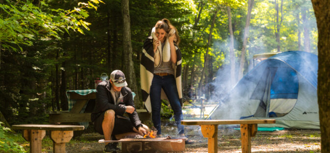 a couple stands over a campfire next to a tent in the woods