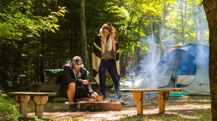 a couple stands over a campfire next to a tent in the woods
